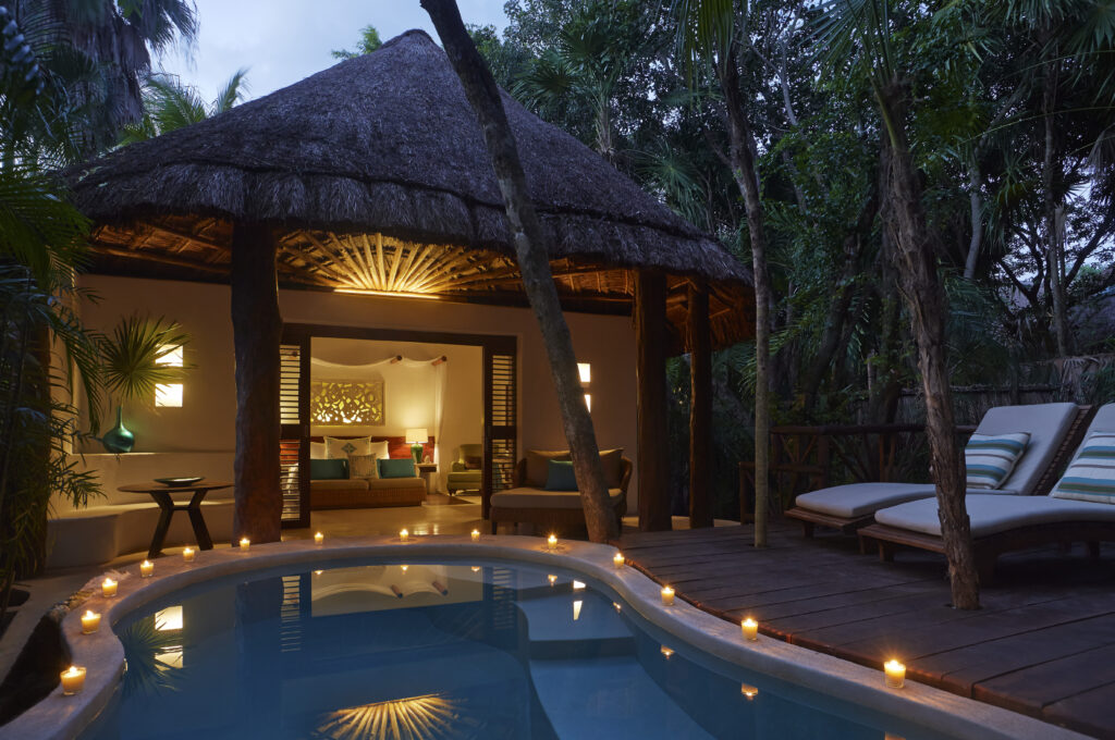 VRM46 - Petróleo y Energía Thatched-roof bungalow at dusk with an illuminated open living area, tropical trees, and a candlelit pool edge.
