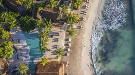 Aerial view of a tropical beach resort with palm trees, thatched roofs, a winding pool, and sun loungers along the sandy shore.