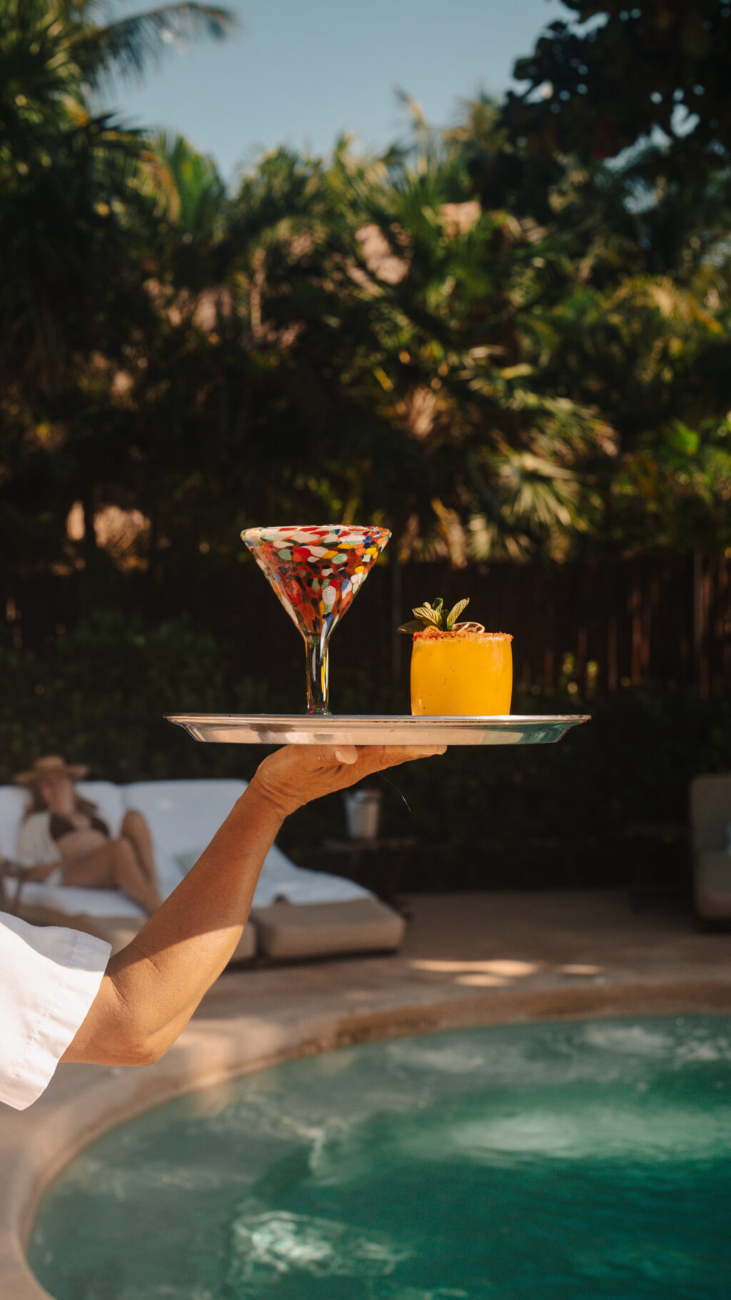 VRM69 - Petróleo y Energía Waiter's hand carries a tray with a colorful mosaic cocktail glass and a bright yellow drink garnished with mint, poolside.