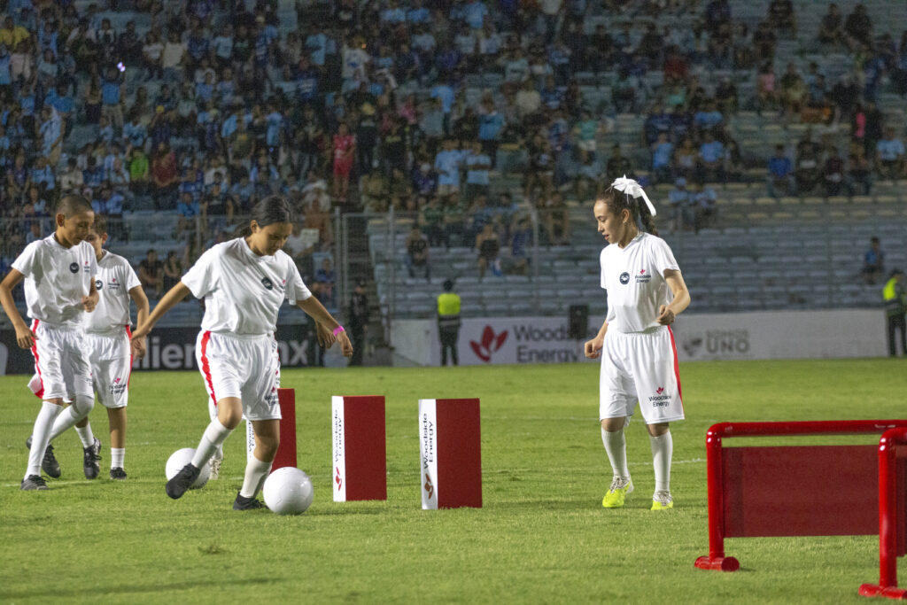 WOODSIDE CAMPEON DE CAMPEONES 2 - Petróleo y Energía Youth soccer players in white jerseys dribble around red obstacle posts on a green field with a packed stadium crowd in the background