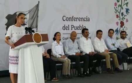 Woman in a white dress speaks at a podium with the Mexican seal; a panel of seven people sits on stage behind her at a 'Conferencia del Pueblo' event, with a decorative mural and floral artwork in the background.