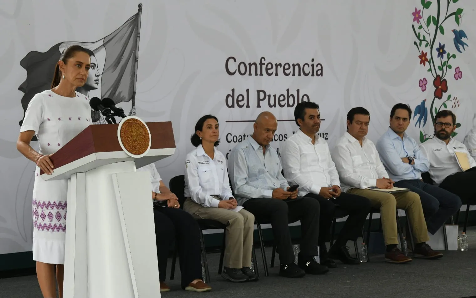 Woman in a white dress speaks at a podium with the Mexican seal; a panel of seven people sits on stage behind her at a 'Conferencia del Pueblo' event, with a decorative mural and floral artwork in the background.
