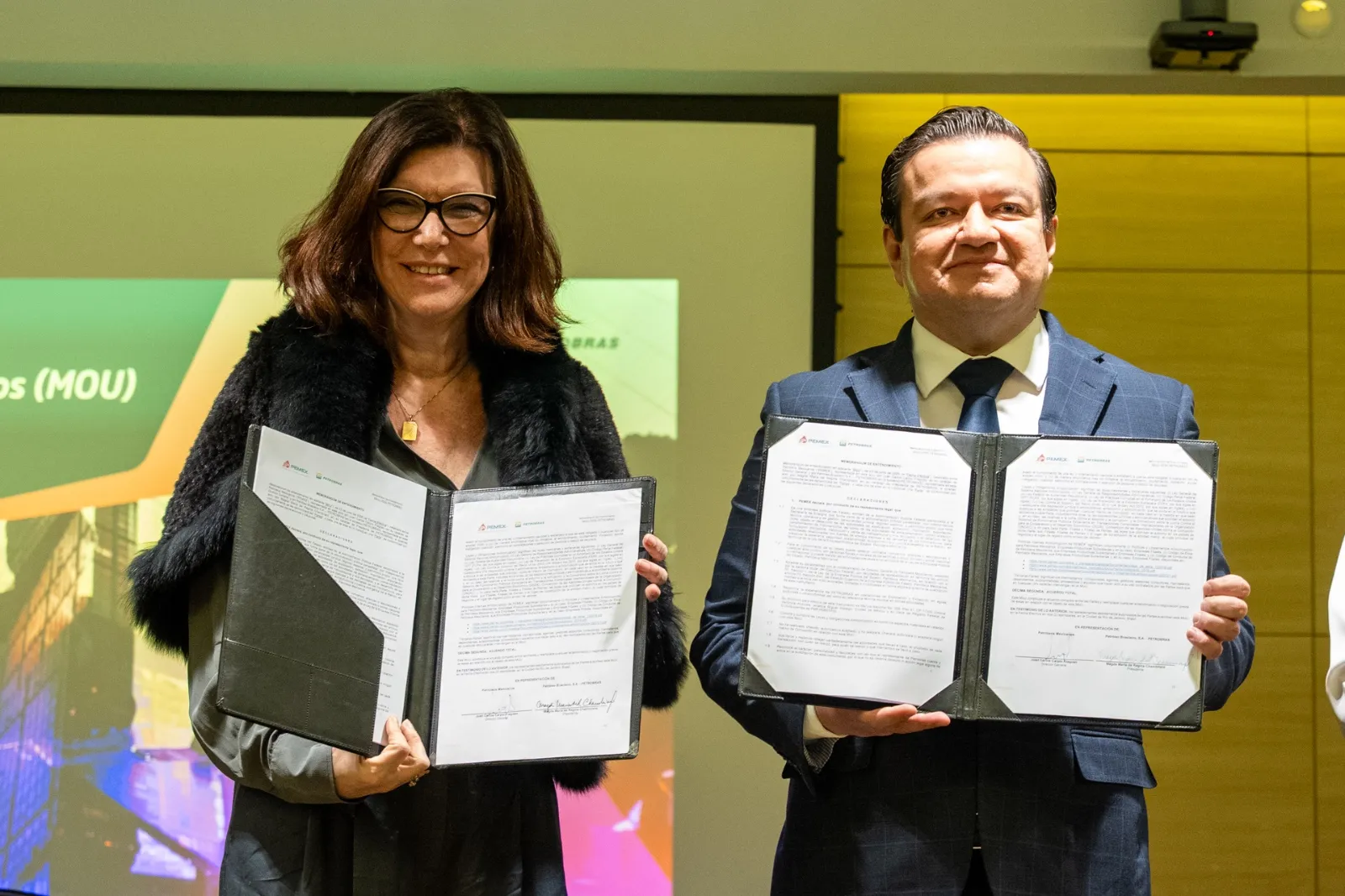 Two professionals in formal attire display signed documents during a signing ceremony, smiling at the camera.