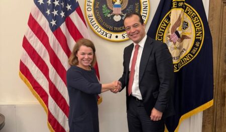 Two professionals shake hands in a formal setting, a woman in a navy dress and a man in a dark suit, with U.S. flags and government seals behind them.