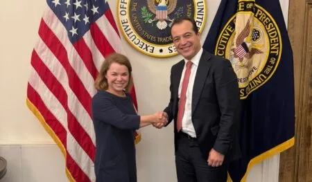 Two professionals shake hands in a formal setting, a woman in a navy dress and a man in a dark suit, with U.S. flags and government seals behind them.