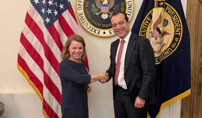 Two professionals shake hands in a formal setting, a woman in a navy dress and a man in a dark suit, with U.S. flags and government seals behind them.
