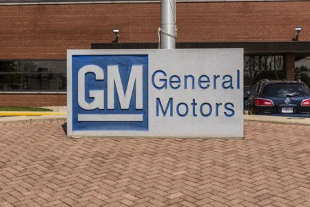 GM General Motors sign on a gray pedestal in front of a brick building with a blue car in the background.