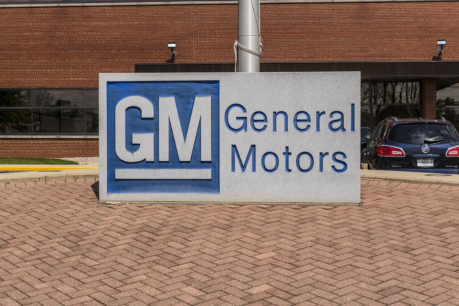 GM General Motors sign on a gray pedestal in front of a brick building with a blue car in the background.