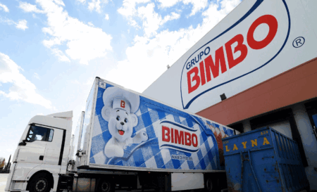 White trucks with large Bimbo logos and a blue refrigerated trailer at a loading dock under a bright sky
