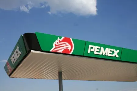 Pemex gas station sign on a green panel with the red logo, against a blue sky.