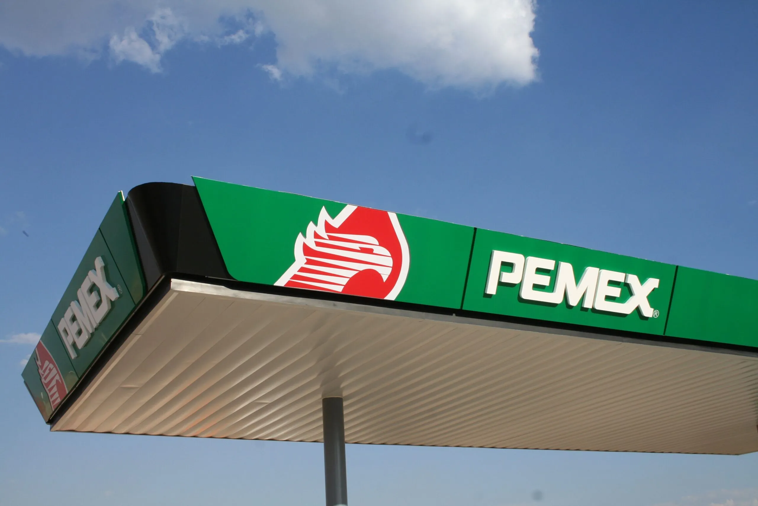 Pemex gas station sign on a green panel with the red logo, against a blue sky.