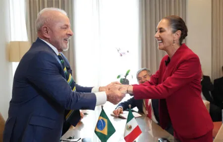 Two dignitaries shake hands across a conference table; Brazil and Mexico flags on the table.