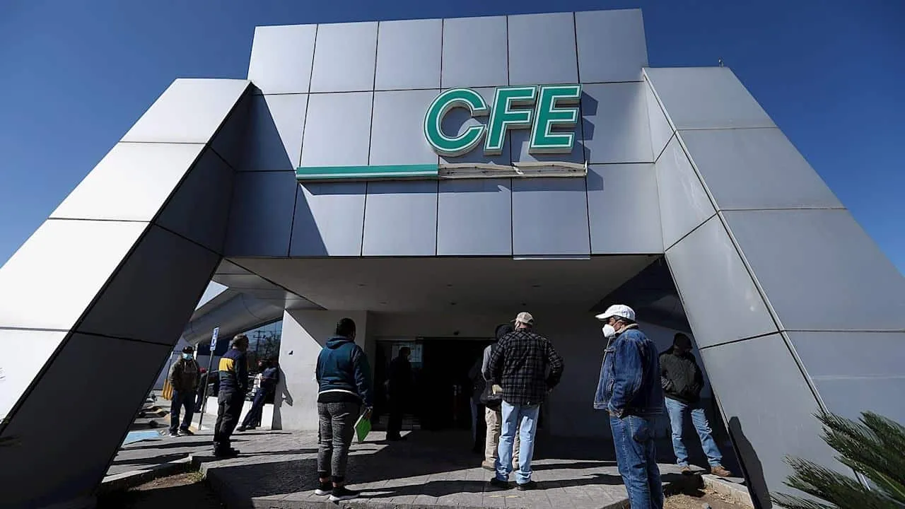 People wearing masks stand in line outside a modern metal building with a large green CFE sign under a clear blue sky.
