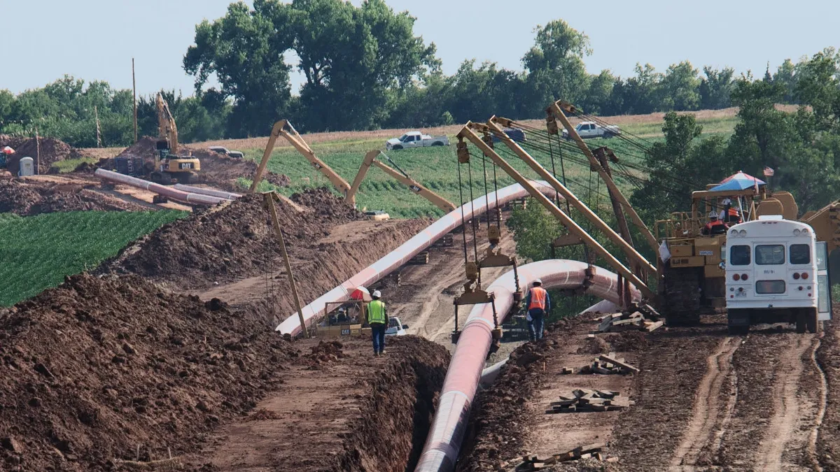 Construction site with large pink industrial pipes laid in trenches and heavy equipment, workers in safety vests guiding pipe sections.