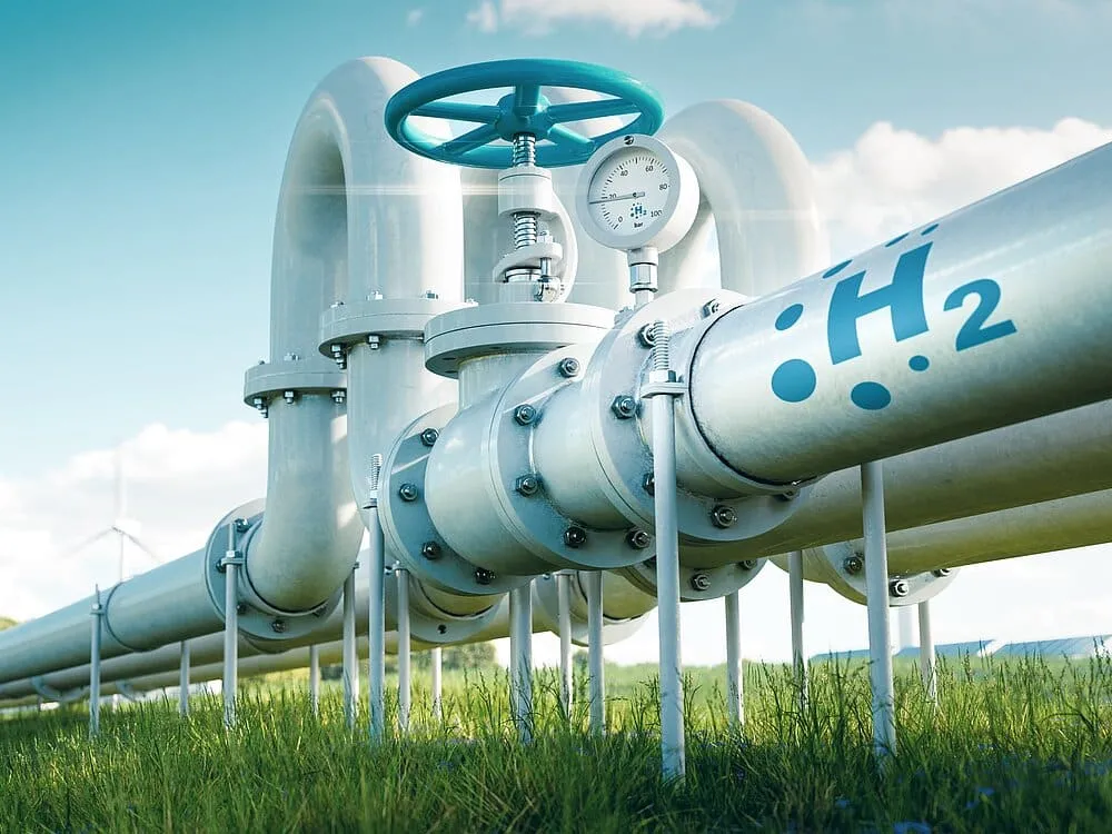 Hydrogen gas pipeline with a large blue-handled valve and pressure gauge outdoors in a grassy field under a blue sky.