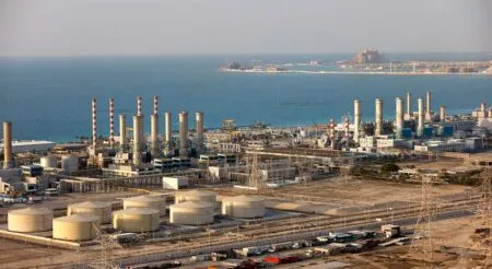 Coastal oil refinery with tall smokestacks, storage tanks, and extensive piping by the sea.