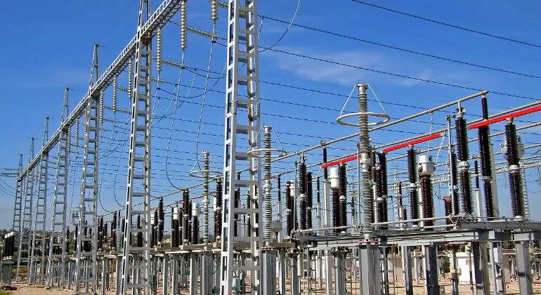 High-voltage substation with metal gantry, insulators, transformers and overhead power lines under a blue sky.