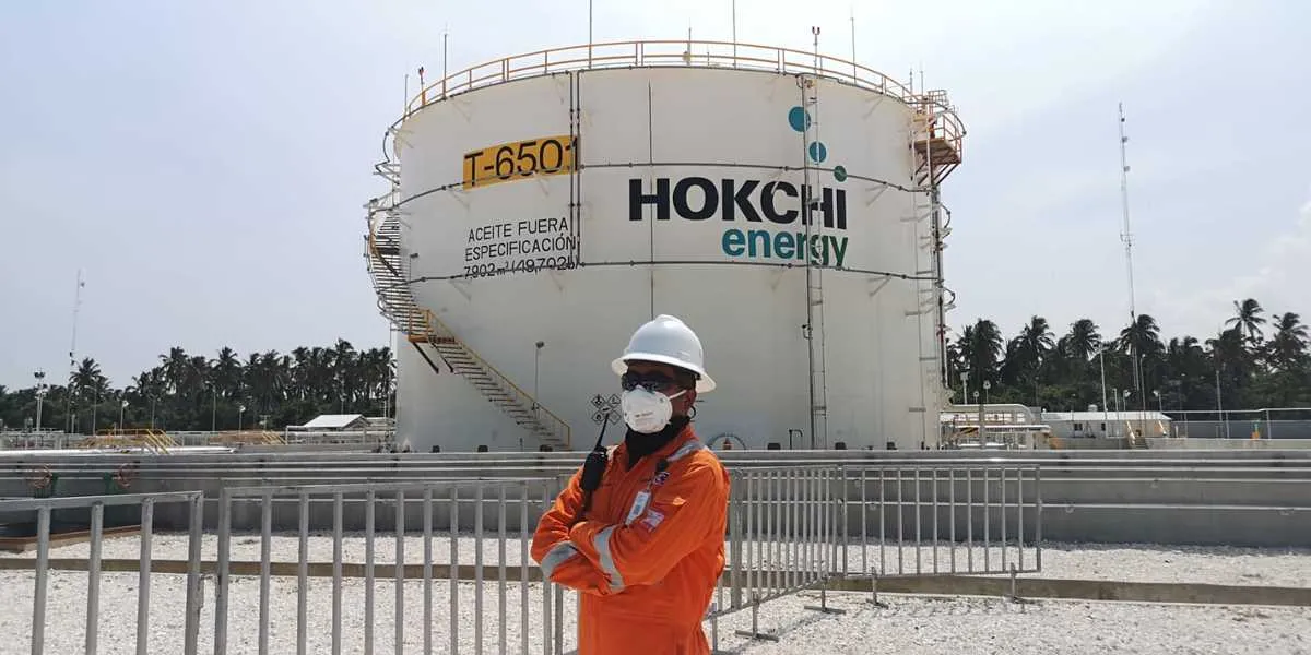 Worker in orange safety suit and white hard hat stands in front of a large Hokchi Energy storage tank.