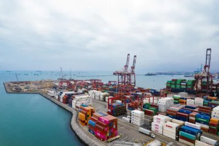 Busy port with stacked shipping containers and cranes along a breakwater by the sea.