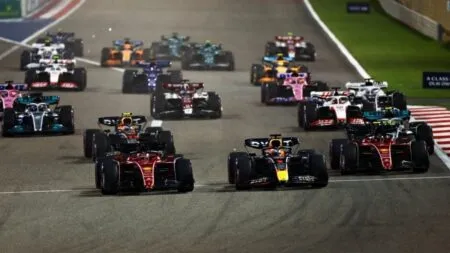 A pack of Formula 1 cars accelerates from the start on a race track, with red Ferraris at the front.