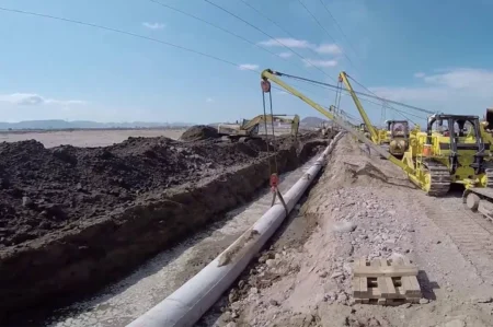 Construction site showing a trench where a large pipeline is being laid, with yellow excavators and rigging nearby under a blue sky.