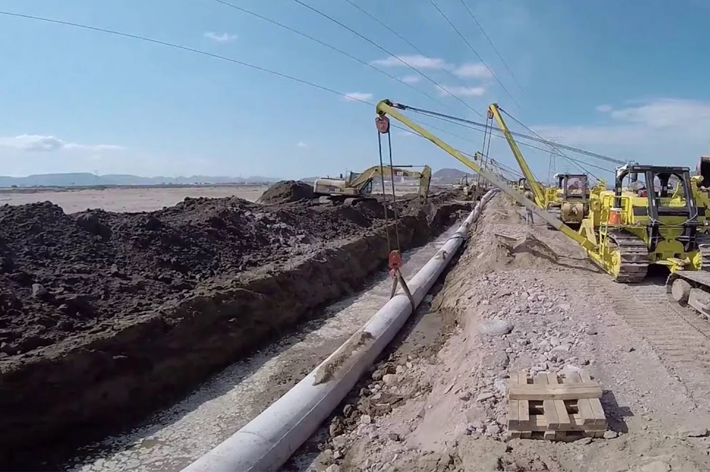 Construction site showing a trench where a large pipeline is being laid, with yellow excavators and rigging nearby under a blue sky.