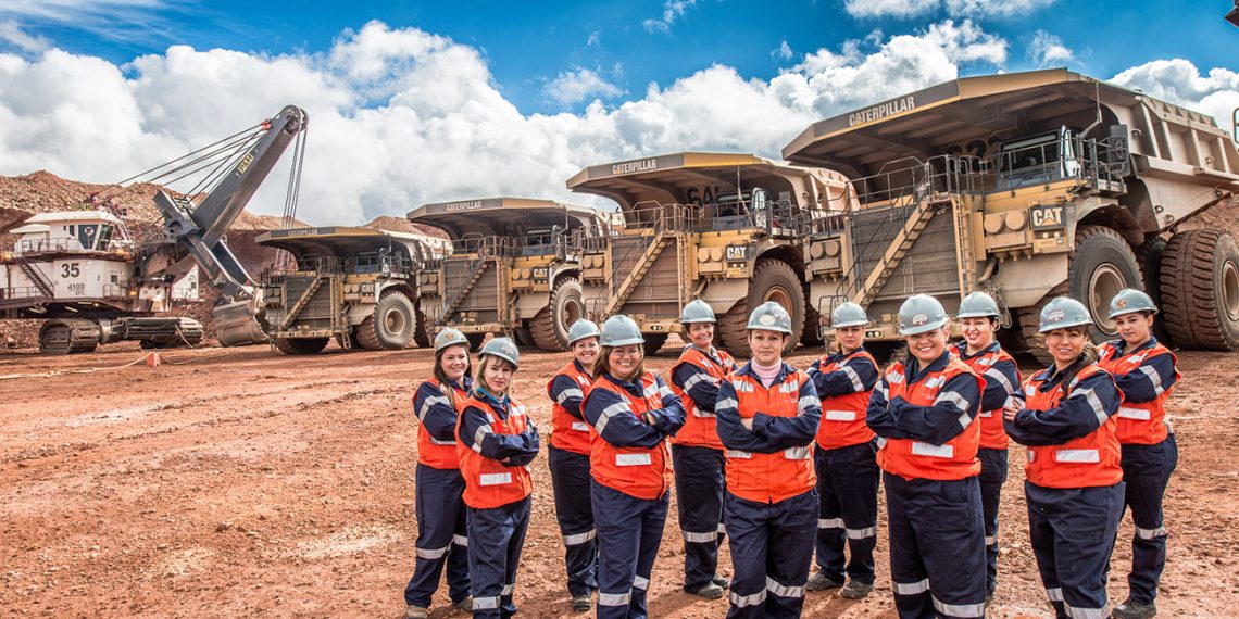 Group of construction workers in orange safety vests and hard hats posing with arms crossed beside enormous mining trucks at a quarry.