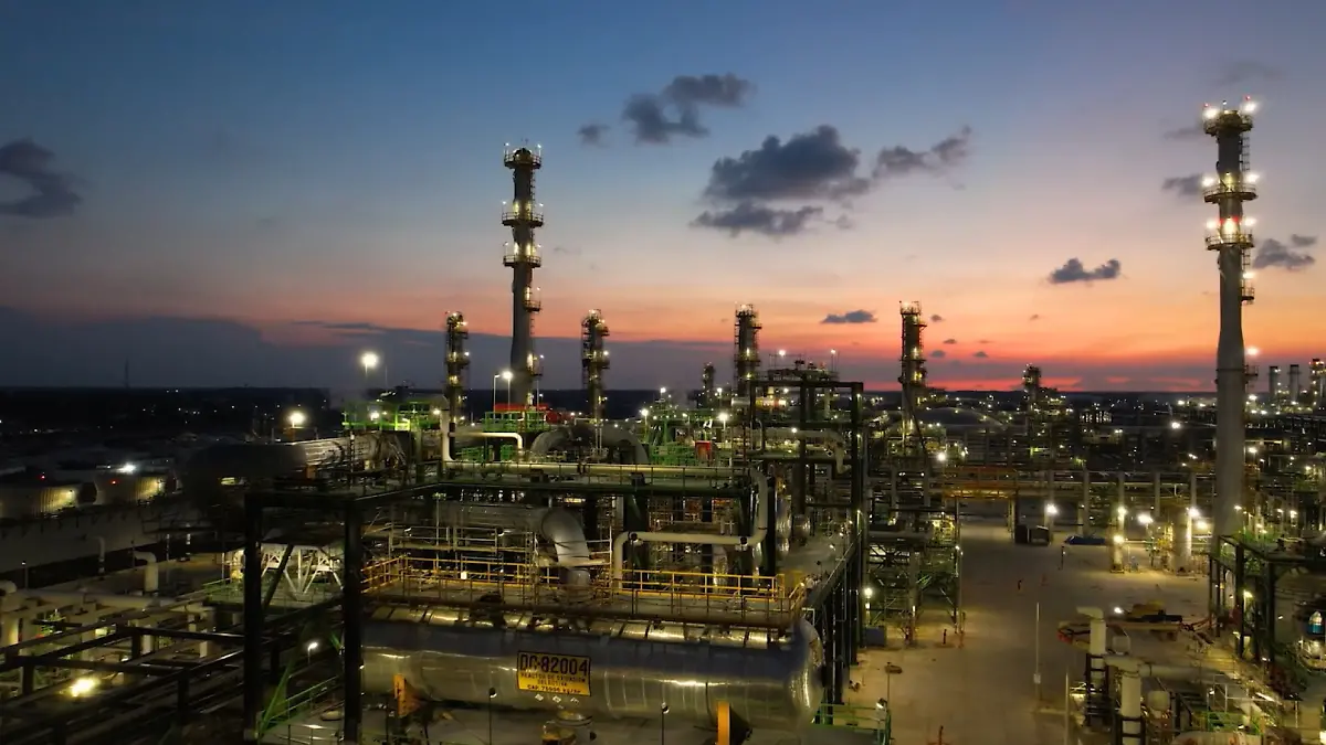 Industrial oil refinery at dusk with tall lit distillation columns and a complex network of pipes and tanks beneath a orange-blue sky at sunset/coastline horizon