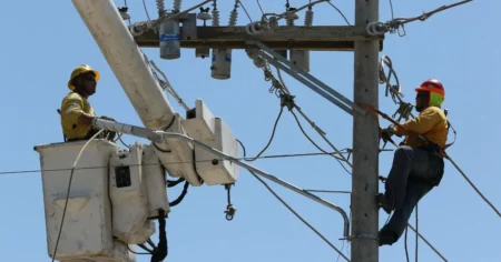 Two utility workers in yellow shirts and hard hats repair power lines from a bucket lift on a utility pole.