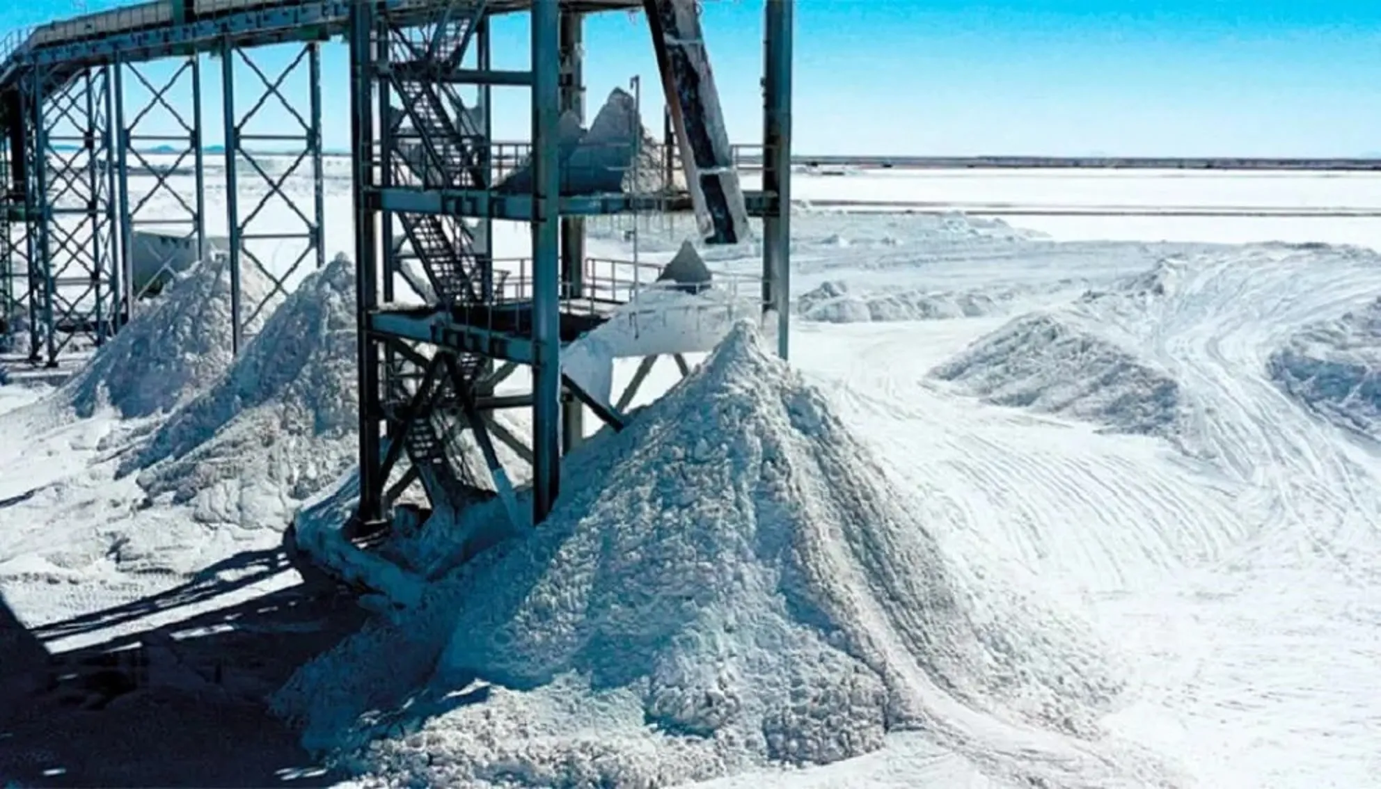 Industrial site with a metal conveyor structure and large piles of white salt in an open yard.