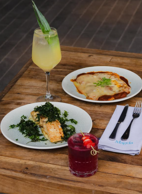 magda -platillo - Petróleo y Energía Two plated dishes and two drinks on a wooden table: herb-crusted salmon with greens, a cheesy baked dish, a yellow pineapple-leaf cocktail, and a red berry cocktail with a strawberry garnish.