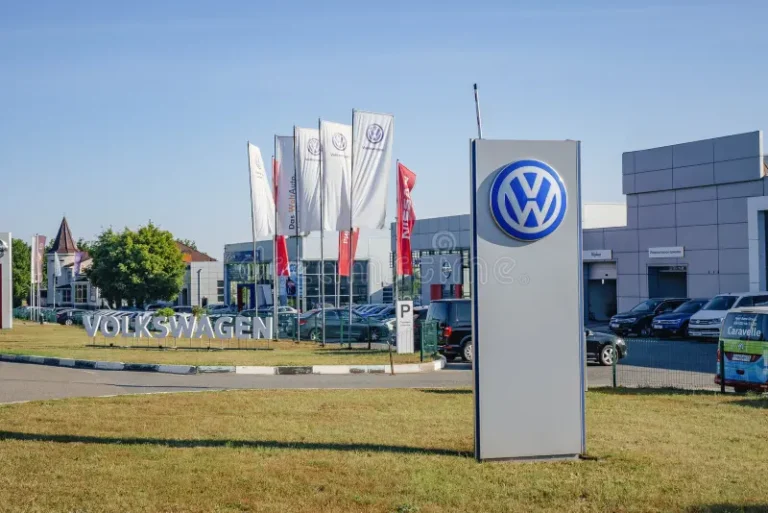 Front view of a Volkswagen dealership with a tall sign featuring the VW logo, flags, and a row of cars in the lot under a clear blue sky