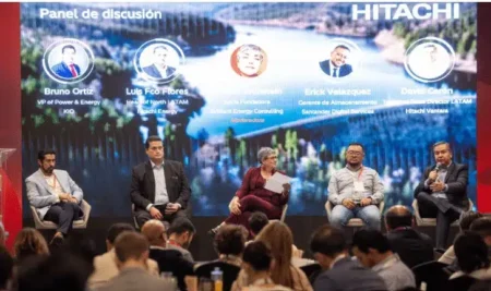 Five panelists sit on a stage in a discussion panel, with an audience in the foreground and a large screen showing portraits and bios behind them (Hitachi branding).