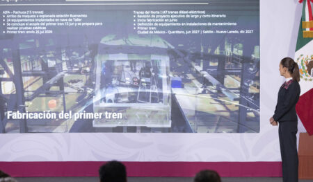 Woman presenting on stage beside a large slide titled 'Fabricación del primer tren' with bullet points visible on screen.