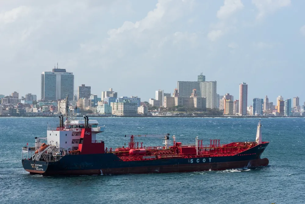 Red and black chemical/tanker ship sailing past a city skyline along the water.