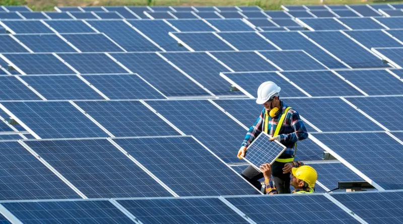 Two workers in hard hats install a solar panel among rows of blue photovoltaic panels in a solar field
