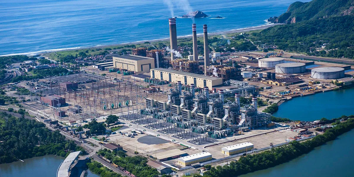 Aerial view of a coastal industrial plant with tall smokestacks, cooling towers, and complex machinery near the shoreline and hills introducing greenery nearby.