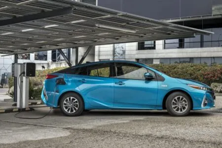 Blue electric sedan plugged into a public charging station under a metal canopy.