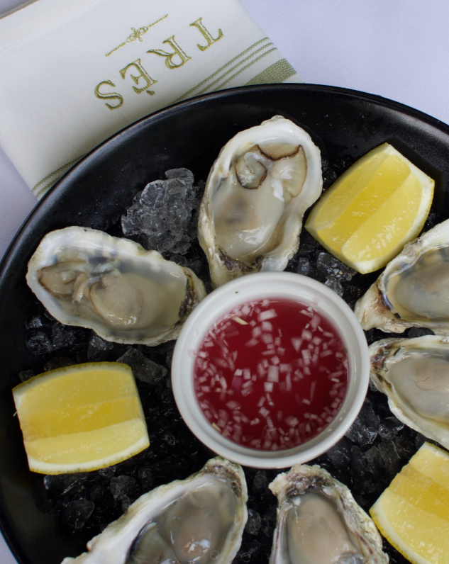 tres1 - Petróleo y Energía Oysters on ice arranged in a circle around a cup of red dipping sauce, with lemon wedges on a black plate.