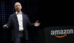 Business presenter in a dark suit speaks on stage beside a black podium with an Amazon logo and orange arrow.