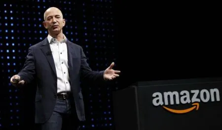 Business presenter in a dark suit speaks on stage beside a black podium with an Amazon logo and orange arrow.