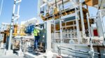 Worker in a hard hat and high-visibility vest operates a control panel amid industrial piping and yellow machinery at an outdoor facility.