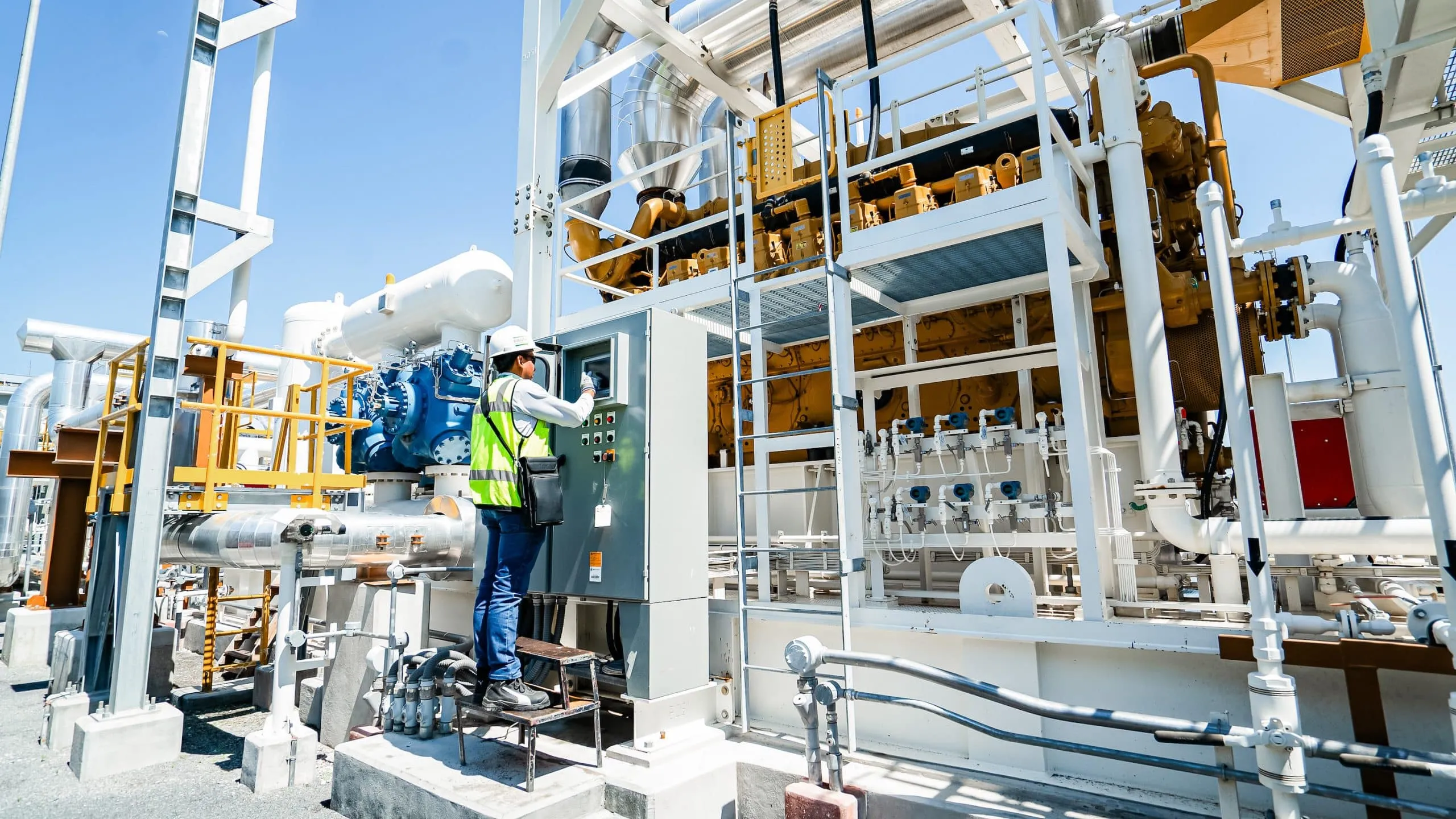 Worker in a hard hat and high-visibility vest operates a control panel amid industrial piping and yellow machinery at an outdoor facility.
