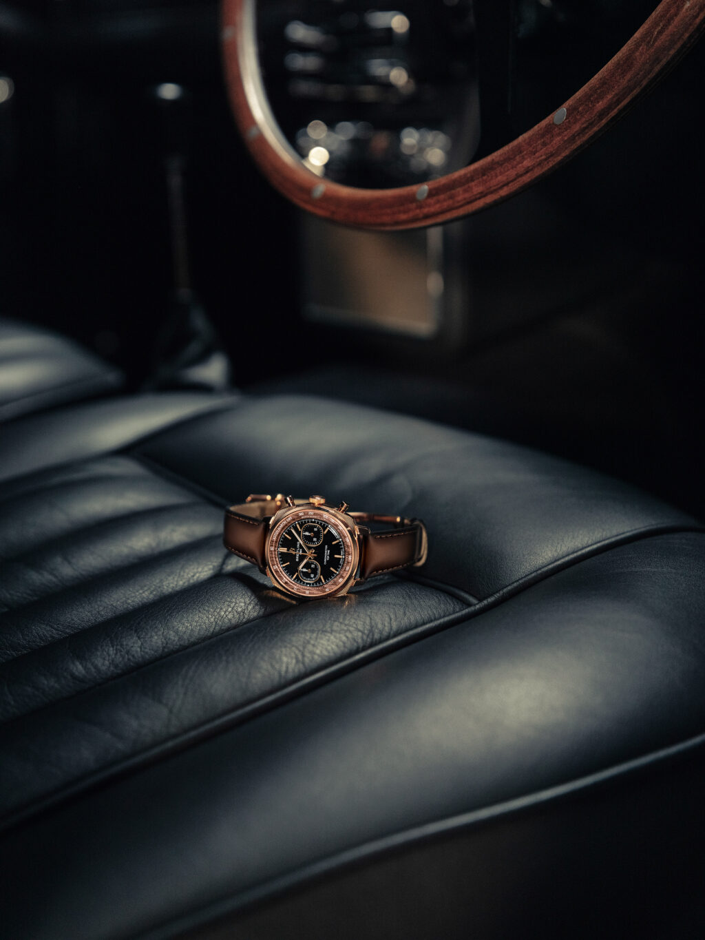 04_Breitling-Top-Time-B01-Chronograph-41-Tribute-to-Aston-Martin-DB5_RB0178131B1X1_RGB - Petróleo y Energía Rose gold wristwatch with a brown leather strap resting on a dark car seat, blurred steering wheel in the background.
