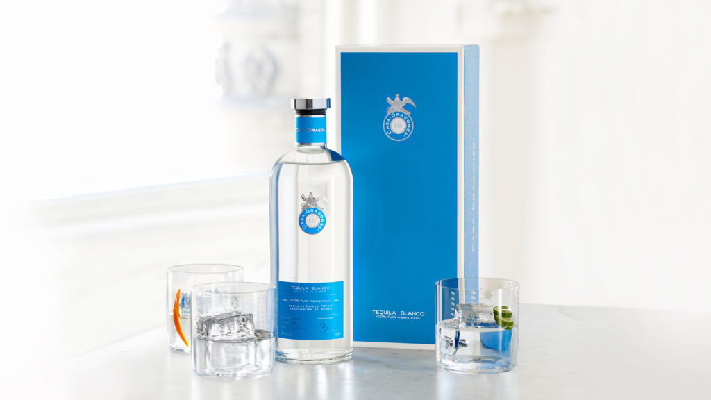 060515_CD_Blanco_Rocks_1-videosize - Petróleo y Energía Bottle of Casa Dragones Tequila Blanco with a blue label beside a matching blue box, two glasses with ice on a white countertop.