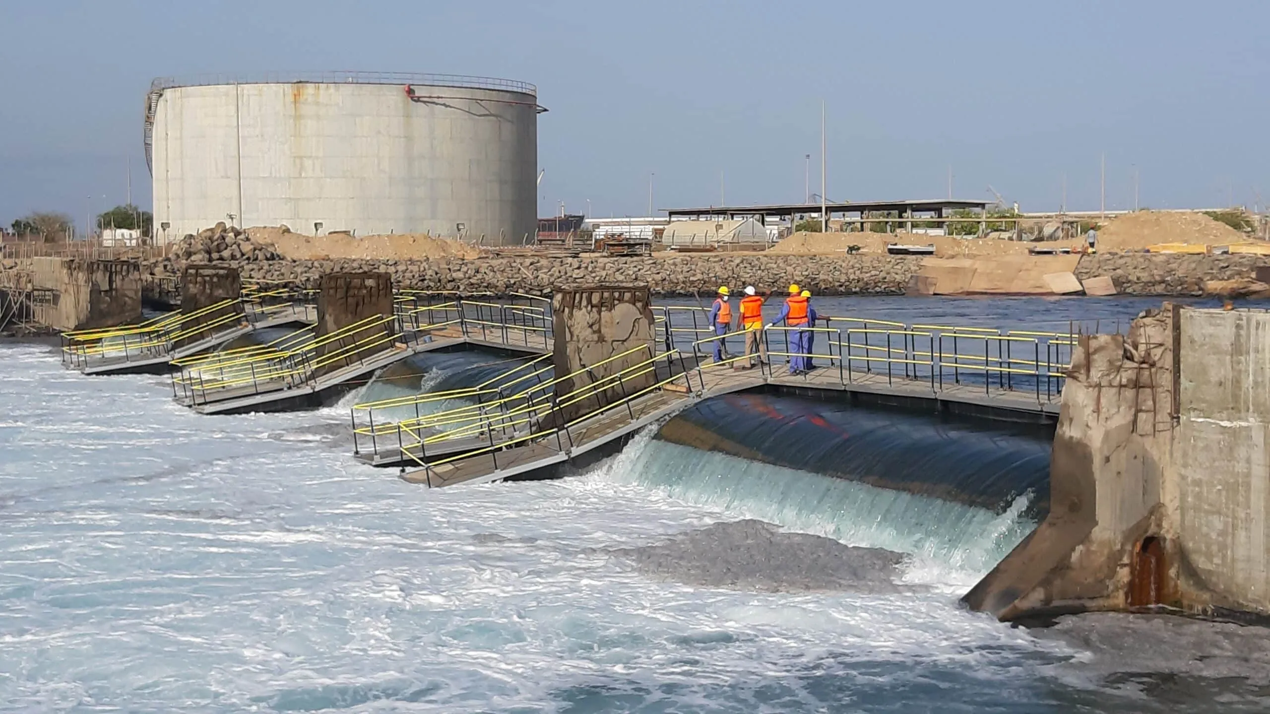 Industrial waterfront construction site with workers in orange vests on a metal walkway beside a large concrete storage tank and seawall.