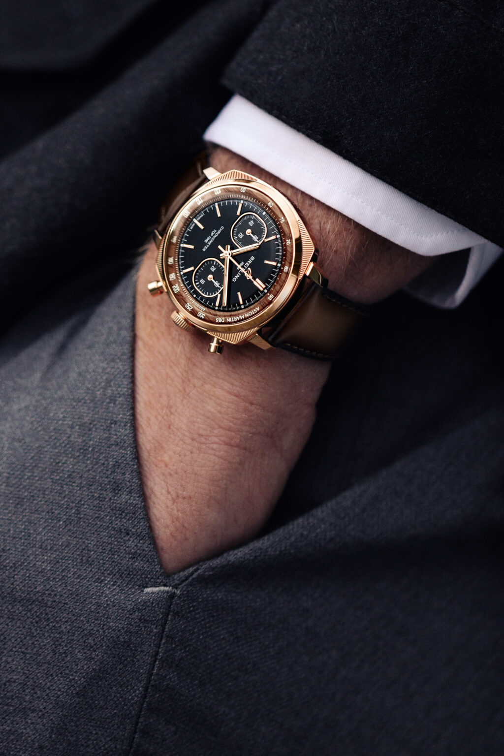 12_Breitling-Top-Time-B01-Chronograph-41-Tribute-to-Aston-Martin-DB5_RB0178131B1X1_RGB - Petróleo y Energía Gold-tone wristwatch with brown leather strap on a wrist in formal attire, close-up view of the watch face and bezel.