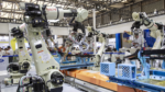 Industrial robotics exhibition with several large robotic arms around a blue conveyor and orange modules.