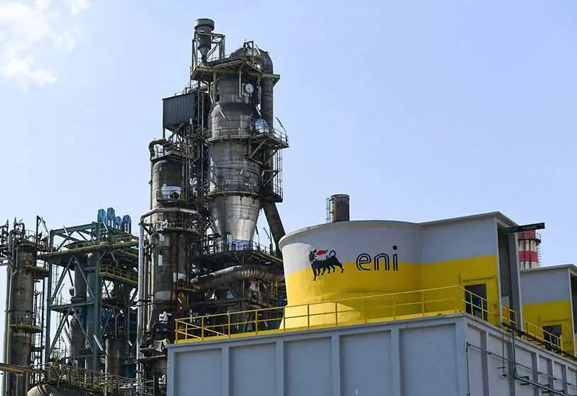 Industrial refinery facility with tall steel towers and piping, featuring a yellow ENI-branded building in the foreground, under a blue sky.