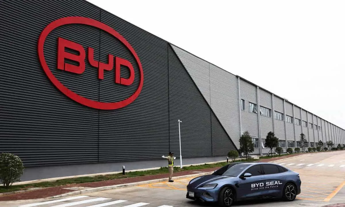Factory exterior with a large red BYD logo on a dark corrugated wall; a BYD Seal electric car is parked in the foreground.